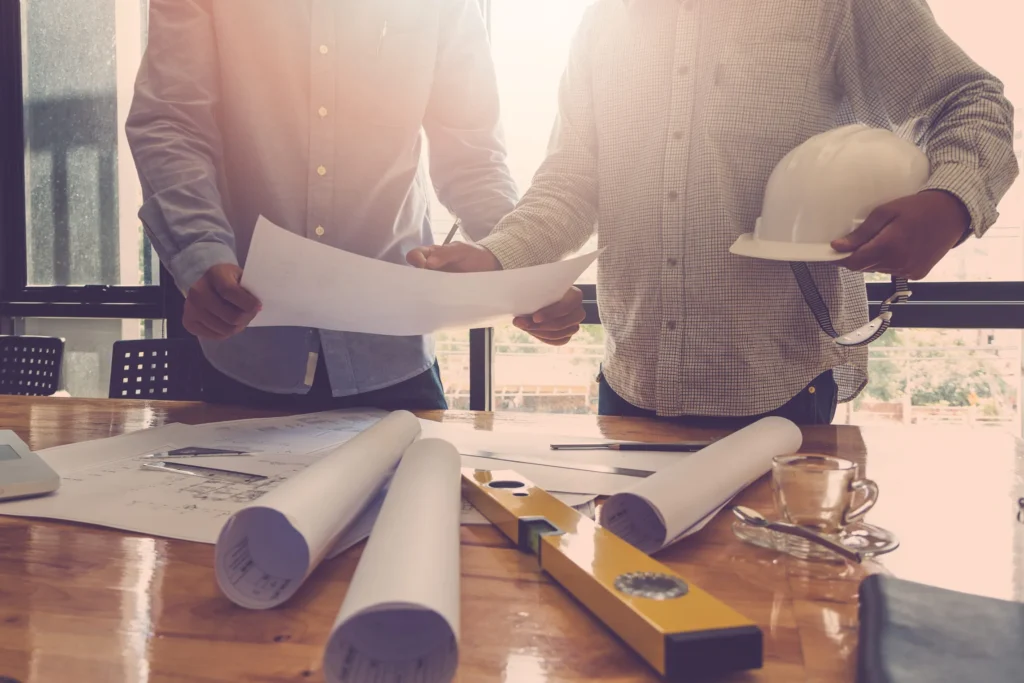 Dos individuos discutiendo sobre planos arquitectónicos y herramientas de ingeniería en una mesa de trabajo.