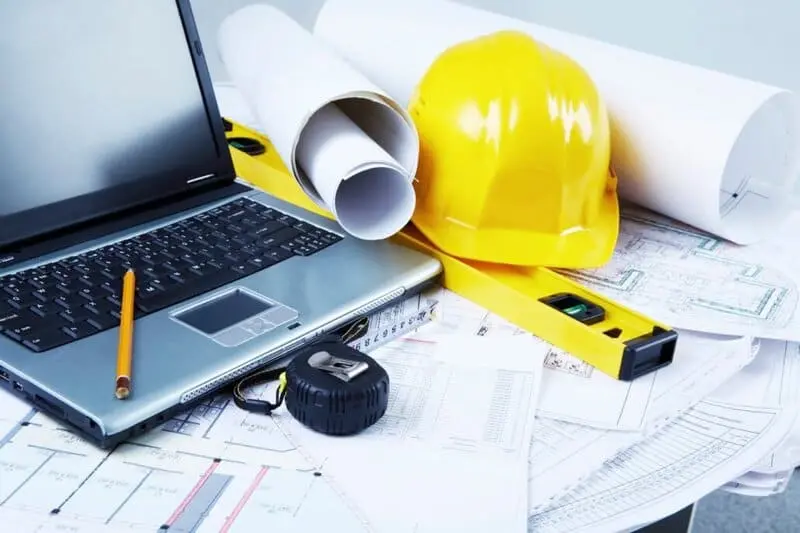Escritorio de trabajo con casco de seguridad, planos arquitectónicos, una laptop y una cinta métrica, representando un ambiente de construcción o ingeniería.