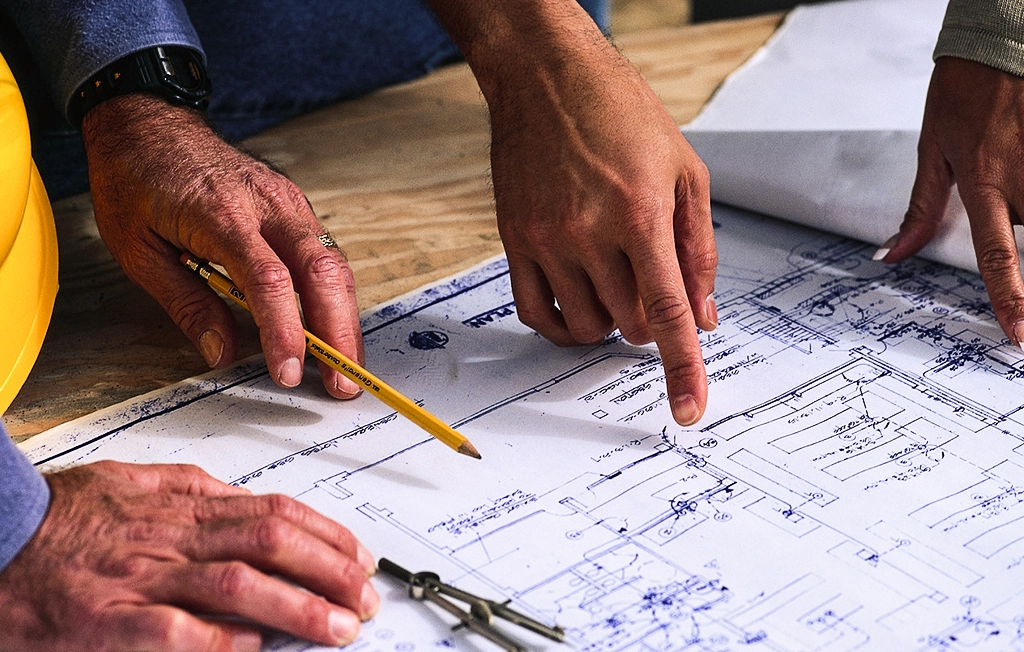 Manos trabajando en planos arquitectónicos con un casco de seguridad y compases visibles.